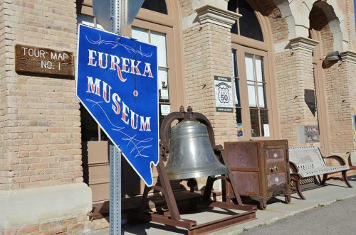 Sign for Eureka Museum in front of a historic building, featuring a large bell and wooden furniture.
