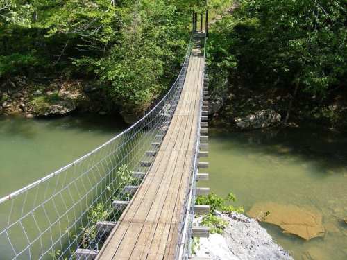 A wooden suspension bridge spans a calm river, surrounded by lush green trees and rocky banks.