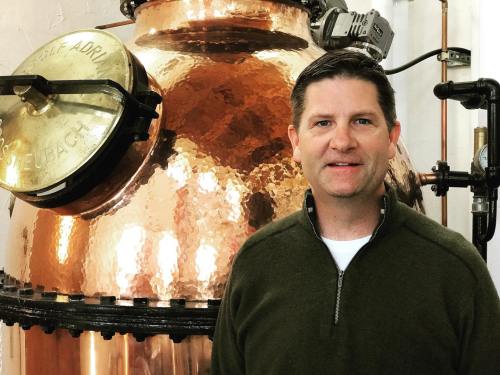 A man stands in front of a large, shiny copper distillation still in a distillery setting.