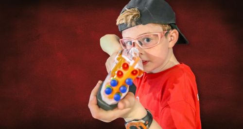 A young boy in a red shirt and cap aims a colorful toy gun, wearing glasses and a focused expression.
