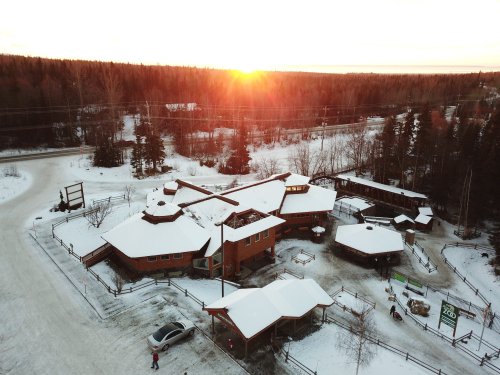 Aerial view of a snow-covered building complex at sunset, surrounded by trees and a road.