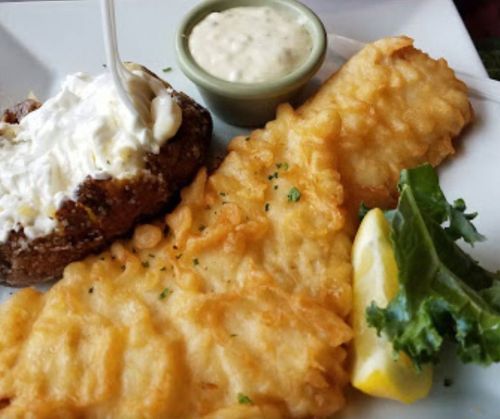 A plate featuring two pieces of golden fried fish, a side of tartar sauce, a lemon wedge, and a baked potato with sour cream.