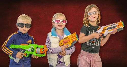 Three children pose with toy blasters, wearing sunglasses and playful expressions against a red background.