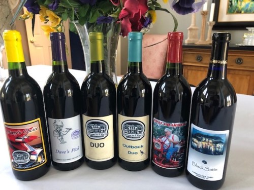 A lineup of seven wine bottles with colorful labels, displayed on a table with flowers in the background.