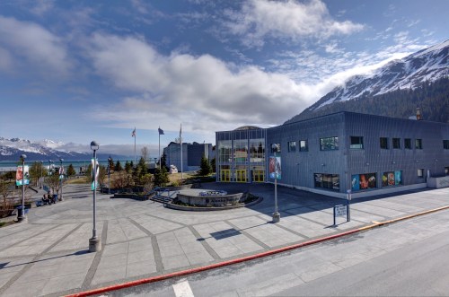 A modern building with large windows, surrounded by trees and mountains, under a partly cloudy sky.