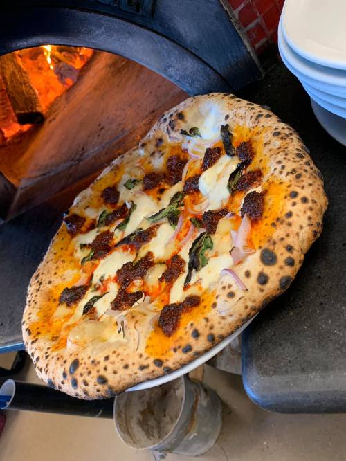 A freshly baked pizza with melted cheese, toppings, and a charred crust, resting near a wood-fired oven.