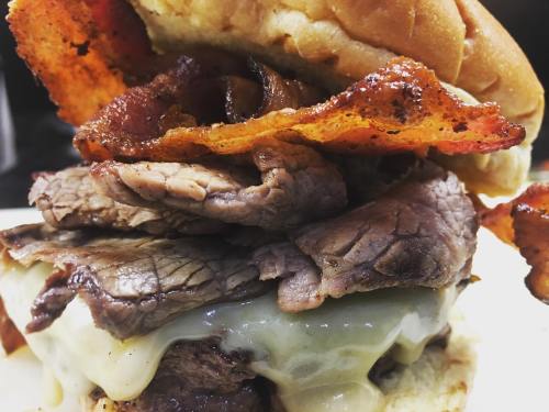 A close-up of a hearty sandwich with layers of beef, melted cheese, and crispy bacon, all in a toasted bun.