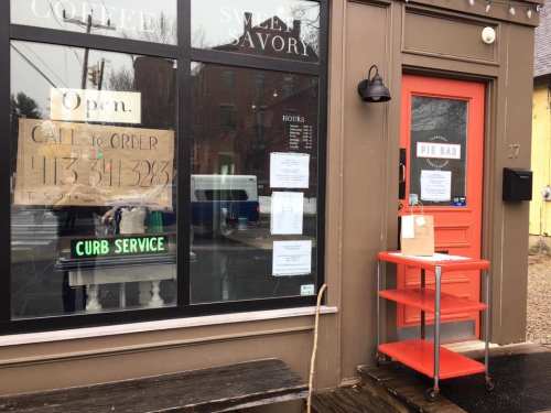 A storefront with a red door and a sign reading "Open" and "Curb Service" in front of a pie bar.