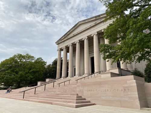 Exterior view of the National Gallery of Art, featuring grand columns and steps, surrounded by trees under a cloudy sky.