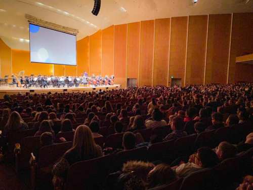A large audience watches an orchestra perform on stage in a spacious concert hall.