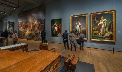 Visitors admire large historical paintings in a museum, with ornate frames and a wooden display in the foreground.