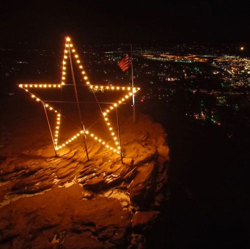 A large illuminated star on a rocky outcrop at night, overlooking a city lit up below. An American flag is nearby.