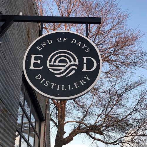 Sign for "End of Days Distillery" featuring a circular logo against a clear sky and tree branches.