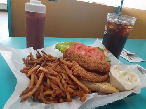A plate of fried fish, fries, lettuce, tomato, a roll, and a cup of soda with a bottle of sauce on a teal table.
