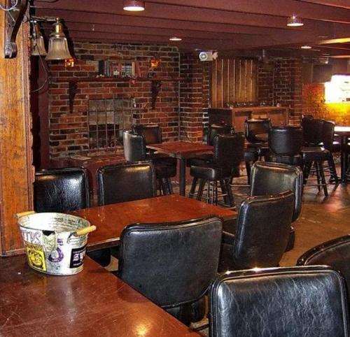A dimly lit bar interior with wooden tables, black leather chairs, and a brick wall featuring a fireplace.
