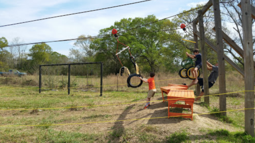 A child plays on a tire swing while another child climbs on a wooden structure in a grassy outdoor area.