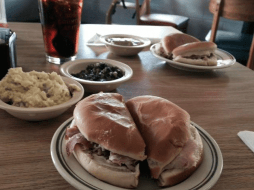 Two sandwiches on a plate, with sides of mashed potatoes, greens, and a drink in a casual dining setting.