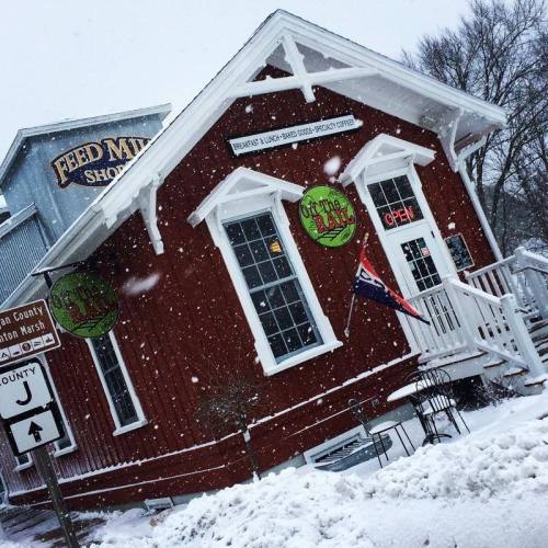 A red building with white trim, featuring "Open" signs, surrounded by falling snow in a winter scene.