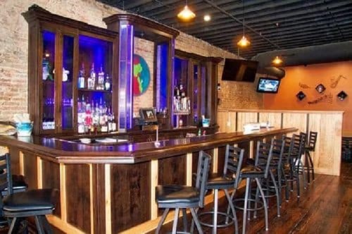 A stylish bar with a wooden counter, illuminated shelves of liquor, and black stools in a cozy setting.