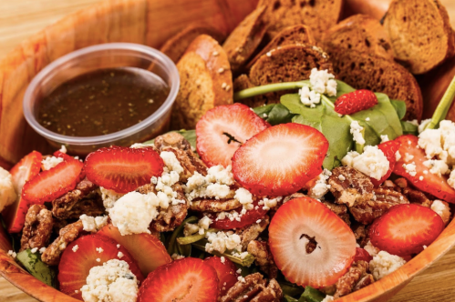 A wooden bowl filled with spinach salad topped with strawberries, pecans, feta cheese, and a side of dressing and bread.