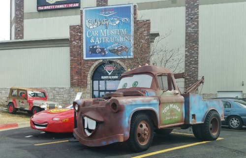 A rusty tow truck and a red race car are parked outside a car museum, with a large sign above them.