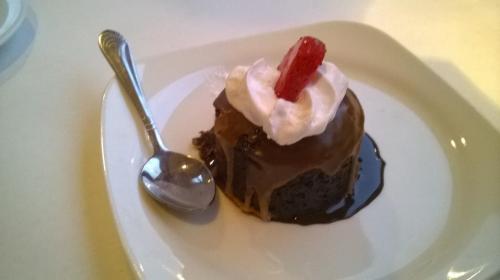 A chocolate dessert topped with whipped cream and a red candy, served on a white plate with a spoon.