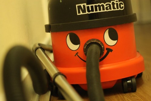 A cheerful red Numatic vacuum cleaner with a smiling face, resting against a wall on a wooden floor.