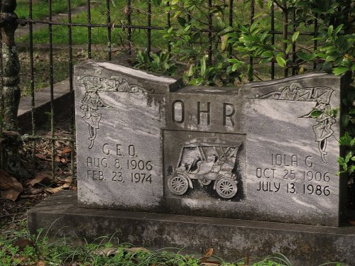 A weathered gravestone with the names "G.E.O." and "Iola G." along with dates and an engraved vintage car design.