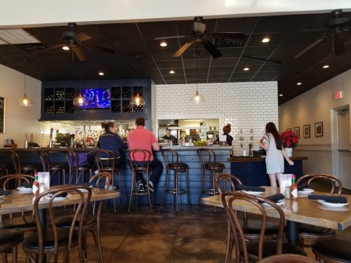 Interior of a restaurant with a bar, patrons seated, and a staff member serving at the counter. Warm, inviting atmosphere.