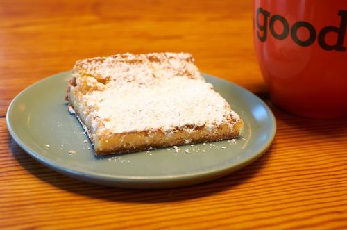 A slice of powdered sugar-dusted dessert on a green plate next to a bright orange mug with the word "good."