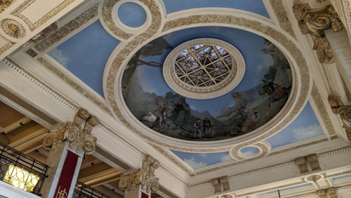 Ornate ceiling with a circular skylight and intricate frescoes, featuring classical figures and decorative moldings.