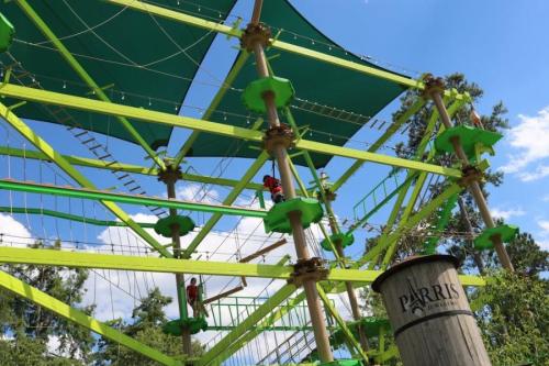 A vibrant green climbing structure with ropes and platforms, set against a blue sky and trees. Two people are climbing.
