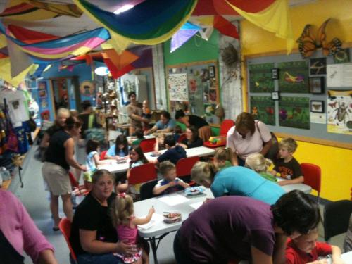 A colorful room filled with children and adults engaged in activities at tables, with decorations hanging from the ceiling.