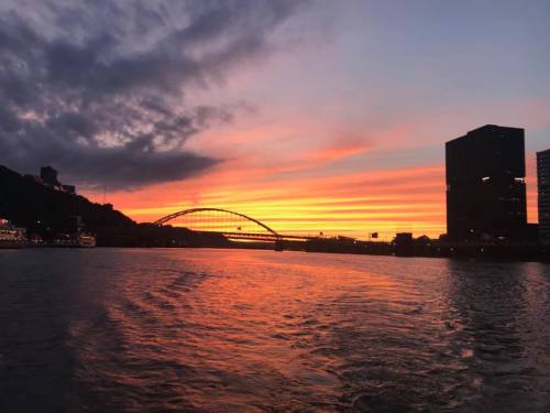 A vibrant sunset over a river, with a bridge and city skyline silhouetted against colorful clouds.