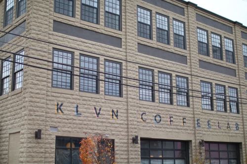 Exterior of a building with large windows and the sign "KLVN COFFEELAB" on the wall, surrounded by power lines.