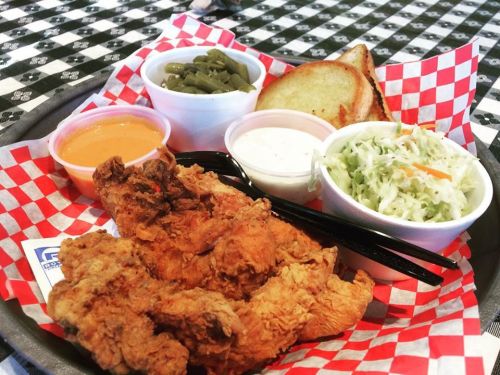 A tray of fried chicken with sides of green beans, coleslaw, a slice of bread, and dipping sauces on a checkered tablecloth.