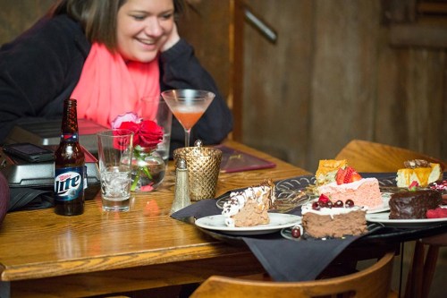 A woman smiles at a table with various desserts, a cocktail, and a beer, set in a cozy restaurant atmosphere.