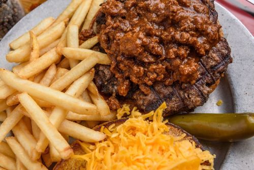 A plate featuring a grilled burger topped with chili, served with fries and shredded cheese, alongside a pickle.