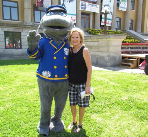 A woman poses with a mascot in a blue uniform outside a building on a sunny day.