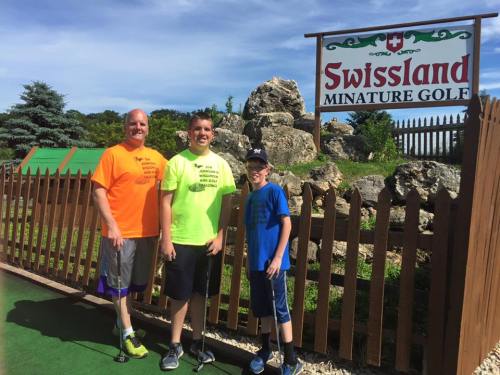 Three people stand in front of a sign for Swissland Miniature Golf, surrounded by greenery and rocks.