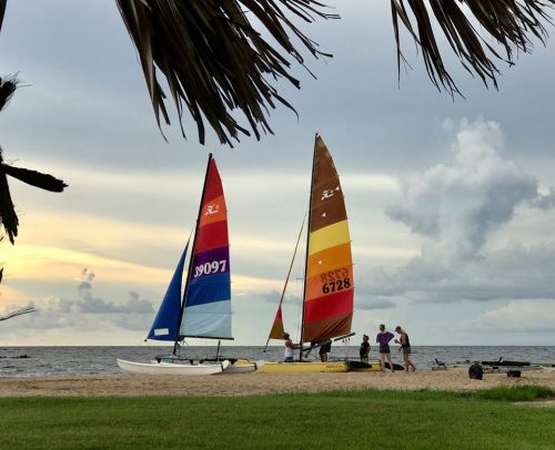 Two colorful sailboats on a beach at sunset, with people preparing to sail under a cloudy sky.
