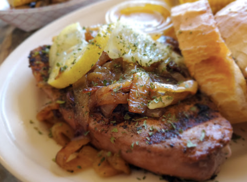 Grilled steak topped with sautéed onions and lemon slices, served with a side of bread and dipping sauce.