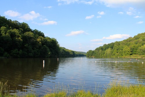 A serene river scene surrounded by lush green hills under a blue sky with scattered clouds.