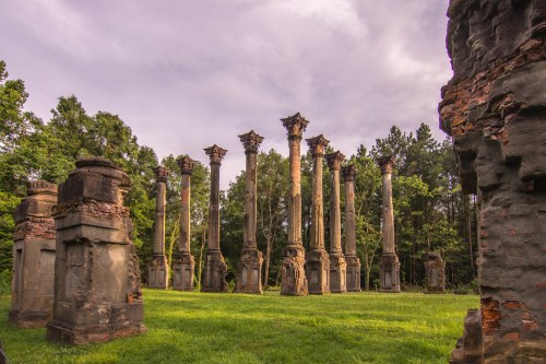 Ruins of tall stone columns surrounded by greenery under a cloudy sky, with remnants of walls nearby.