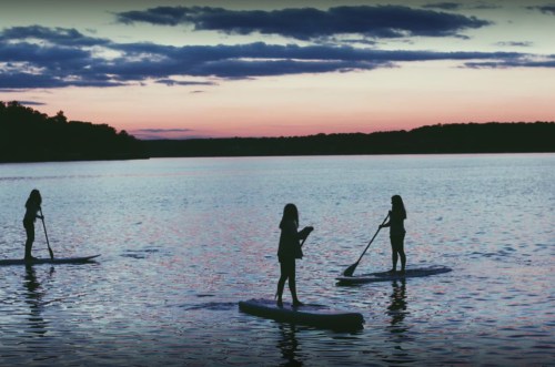 Three people paddleboarding on a calm lake at sunset, with colorful clouds reflecting on the water.