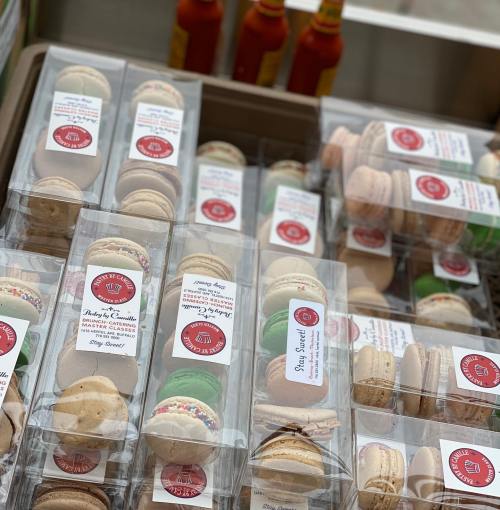 A display of assorted macarons in clear packaging, with bottles of hot sauce in the background.
