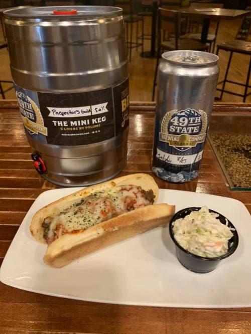A mini keg of beer next to a hot dog topped with cheese and sauce, served with a side of coleslaw on a plate.