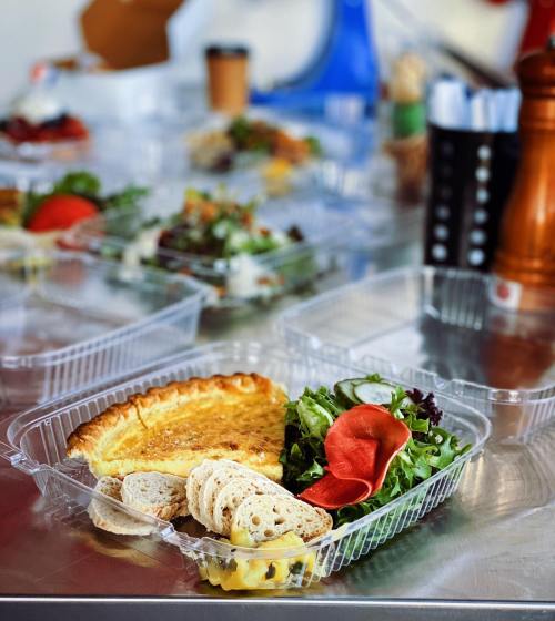 A clear takeout container with a slice of quiche, salad, and slices of bread on a table with other food items in the background.