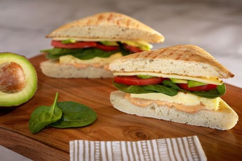 A sandwich with layers of spinach, tomato, and egg on toasted bread, served on a wooden board with an avocado.