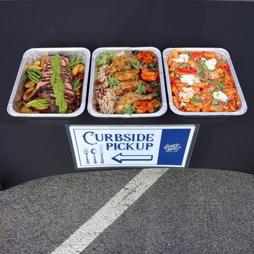 Three trays of food: grilled meats, vegetables, rice, and pasta, with a sign for curbside pickup.